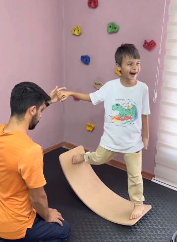 Eğitmen desteğiyle denge tahtasında duran çocuk